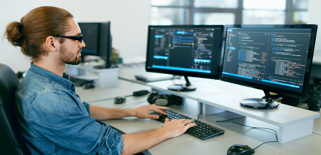 ein junger Mann sitzt am Computer vor zwei Monitoren und programmiert