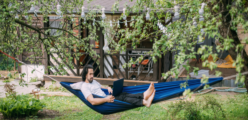 Ein Mann liegt im Garten in einer Hängematte und hat einen Laptop auf dem Schoß.